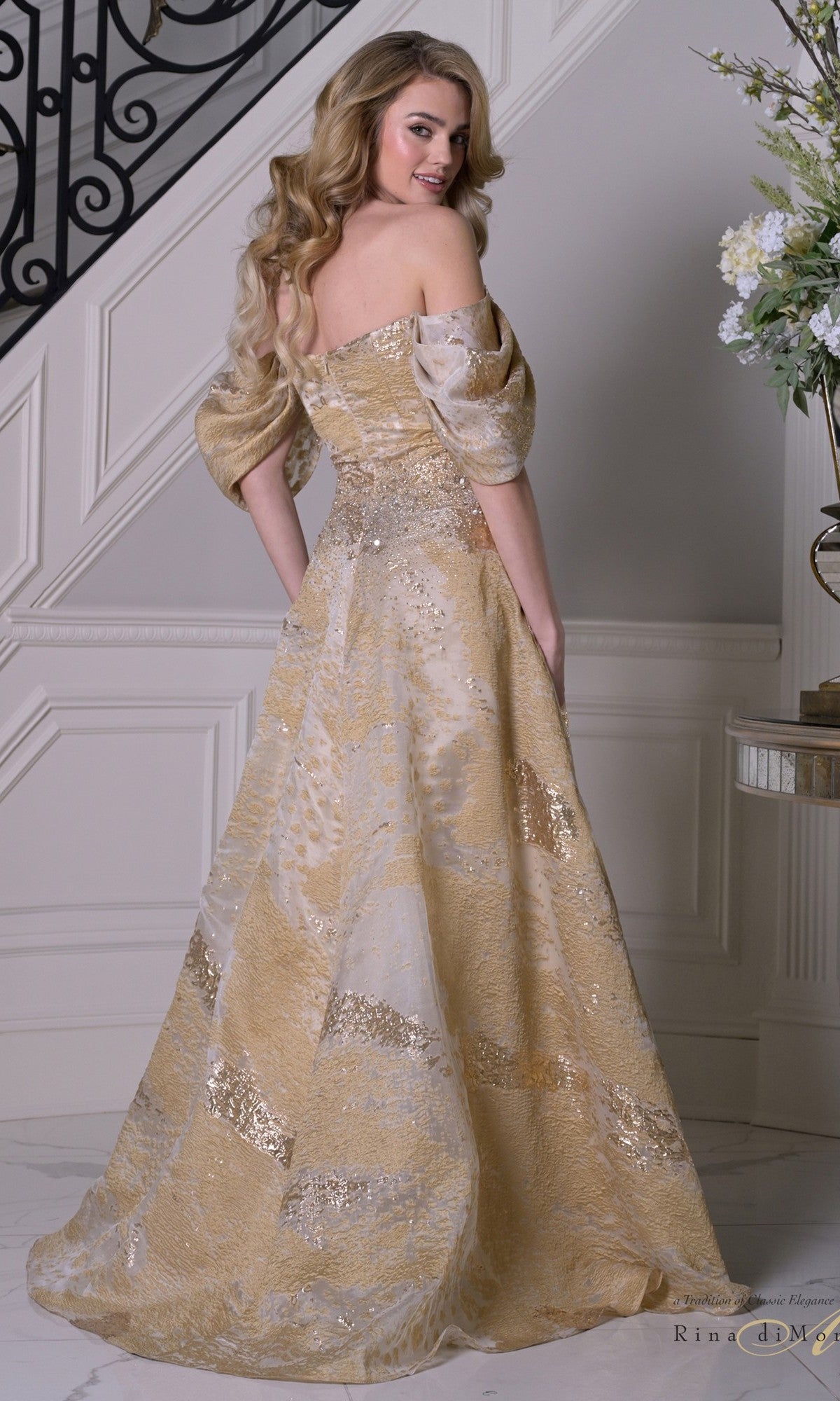 A woman with long blonde hair wears the Rina Di Montella RD3405, an off-the-shoulder gold organza gown with sequins and a beaded bodice, standing by a staircase on a marble floor near a white paneled wall.